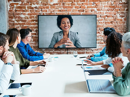 Six men and women sitting at a conference table facing a video screen of woman talking