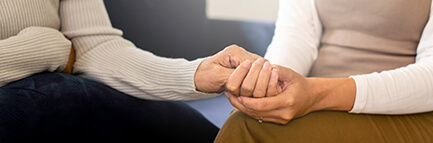 Two people sitting close together and clasping hands to show support and care