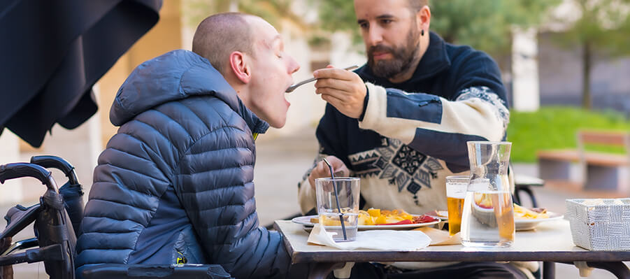 Disabled person eating outside at a restaurant with the help of a caregiver.