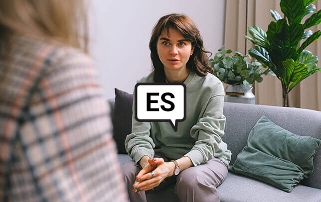Young woman sitting on a couch speaking with a counselor. Letters ES over image to indicate Spanish language